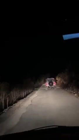 The video captures a night scene on a dirt road with sparse vegetation on the sides. A red SUV is visible driving away from the camera on the road. In the distance to the left, there appears to be a glow of fire, indicating a potential wildfire. The camera is positioned from inside another vehicle, suggesting the person recording is also driving. The overall atmosphere is dark and somewhat eerie due to the nighttime setting and the presence of fire.