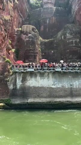 From a boat on a green river, a full shot reveals the Leshan Giant Buddha carved into a red cliff face, partially covered in moss. A crowded platform is constructed at the Buddha's feet. People, some holding red umbrellas, gather along the platform's railing. Red stairs ascend the cliff face to the left of the Buddha's hand. Trees stand on top of the cliff, above the Buddha's head. The sky is bright.