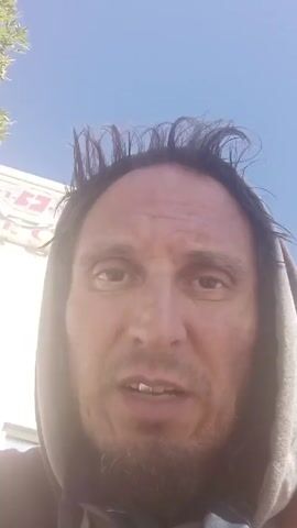 A medium close-up shot focuses on a man's face. He has dark hair that sticks up in places. He's wearing a grey hooded sweatshirt. He has a dark complexion and light brown eyes. He speaks directly to the camera. A clear blue sky is visible in the background. A blurry sign is visible in the upper left of the frame. The lighting is bright.