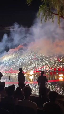 At night, a crowd watches a stage production over water, lit with red lights, that includes bursts of flames. Two people stand in the water holding poles. Smoke hangs in the air. One person in the crowd holds up a phone to record the show. A tree hangs in the upper-right.