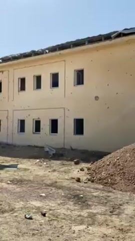 The video shows a damaged two-story building with numerous bullet holes. The windows are missing or broken. The roof appears damaged, and debris is scattered around the building. The surrounding area is barren and dusty. There is a low wall in the distance, suggesting an enclosure or compound. The sky is clear and blue. There are no people visible in the video.
