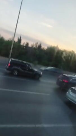 The video shows a view from a moving vehicle on a highway. The sky is visible with some clouds, and the sun appears to be setting or rising, casting a warm light. A dense line of trees runs along the side of the highway. Several cars are seen on the highway, moving in the same direction. These include a black SUV, a silver Toyota Prius, a white sedan, a dark-colored sedan, and a white van. The cars are in motion, creating a blurred effect. The road appears to be wet, suggesting recent rain. The camera is positioned to capture the side view of the traffic flow.