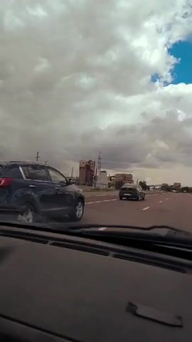 The video shows a view from inside a vehicle driving on a highway. Several other vehicles are visible on the road, including a dark-colored SUV in the left lane and a black car further ahead in the right lane. The sky is filled with large, dramatic clouds, with patches of blue sky visible. In the distance, there are buildings and power lines. The camera perspective suggests the footage is captured from the driver's or passenger's seat, looking forward through the windshield. The road appears to be asphalt and has white lane markings. The overall scene is one of travel on a cloudy day.
