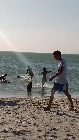 A group of people are at a beach.  Some people are wading in the water. Others are walking along the beach. The beach is sandy. The water is light blue. The sky is bright. There is a glare from the sun.  The people are wearing casual beach attire.