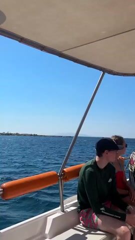The video shows a group of people sitting on a boat on a sunny day. The boat is moving through blue water with a clear blue sky above. The people are sitting under a canopy that provides shade. Some people are looking out at the water, while others are talking to each other. There are a few bags on the boat, including a straw bag and a brown paper bag. The people appear to be on a pleasure cruise.
