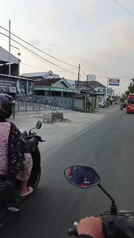 The video shows a first-person view of a person riding a motorcycle down a paved road. On the left side of the road, there are buildings, fences, and parked vehicles. One building has a sign that says "Indomaret." On the right side of the road, there are more buildings, parked cars, and a red bus. The sky is overcast. The rider's hand is visible on the handlebars. The video is taken during the day.