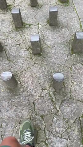 A first-person perspective video shows a person walking on a stone path featuring raised concrete stepping stones. The path is constructed of cracked grey stones, some of which have moss growing in the crevices. The stepping stones are a uniform grey color and are either rectangular or cylindrical in shape. The person is wearing green athletic shoes with white laces, and green shorts. The camera moves as the person steps from one stone to the next.