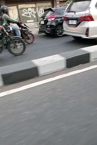 The video shows a street scene. A silver car is parked on the left side of the road. A gray barrier with black and white stripes separates the road from a sidewalk. A person wearing a helmet and a brown jacket is riding a motorcycle. The motorcycle has red shocks and a silver exhaust pipe. The rider is wearing blue jeans and sandals. There are other motorcycles and cars in the background, as well as buildings and billboards. The video appears to be shot from another vehicle moving alongside the motorcycle.