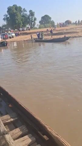 The video shows a muddy river with several wooden boats. On the opposite bank, a large group of people has gathered near some cars and a possible makeshift dwelling.  A few people are near one of the boats on the bank, while a man sits on one of the boats in the foreground.  The water is murky brown. The sky is clear and the sun is shining.