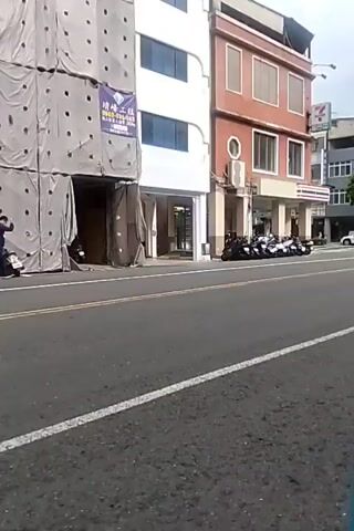 The video opens with a shot of a street in front of a row of buildings. The building on the left is under construction, covered in scaffolding. The middle building is a multi-story structure painted red with white trim. A 7-Eleven convenience store is visible on the ground floor of the red building. A row of motorcycles is parked in front of the 7-Eleven. The building on the far right is white with multiple floors and windows. The road in front of the buildings is asphalt with white lane markings.

In the second half of the video, a person on a black motorcycle rides across the frame from right to left. The rider is wearing a grey t-shirt and a black helmet. The motorcycle has a license plate. The perspective shifts slightly, providing a clearer view of the motorcycles parked in front of the 7-Eleven and the surrounding buildings. A white van is visible parked further down the street on the right. The sky is overcast.