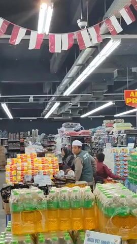 The video shows the interior of a well-lit supermarket. In the foreground, stacked cases of bottled beverages are visible. In the mid-ground, a small group of people are gathered around a display of products. Two individuals, a woman wearing a hijab and a man wearing a white cap and a vest with "ASIFFA" printed on it, are interacting. Behind them, more stacks of products are organized on shelves, and other shoppers can be seen in the background. Overhead, fluorescent lights illuminate the space, and a string of red and white flags is hung across the ceiling, possibly indicating a national celebration. A security camera is also visible mounted on the ceiling.