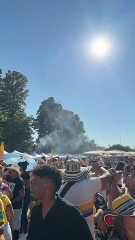 This is a vertical video of an outdoor festival. The sun is high in a clear blue sky, casting a bright glare. An airplane is visible flying high above the trees and a plume of smoke rising from the ground. In the foreground, a crowd of people is gathered. Many are wearing traditional Colombian hats, some of which are black and white striped, others with colorful bands. Some people are wearing yellow shirts with blue and red stripes. There are white tents set up in the background, suggesting vendor stalls or booths. The overall atmosphere appears to be festive and crowded.