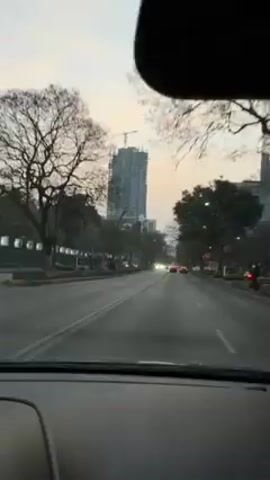 The video shows a view from inside a car driving down a city street. The car is traveling on a multi-lane road with trees lining both sides. In the distance, a tall building under construction can be seen. Other vehicles are on the road, moving in the same direction. The sky is overcast with a hint of color from the setting sun. The car's dashboard is visible, and the speedometer indicates the car is moving at approximately 60 kilometers per hour. A hand briefly appears to adjust something on the dashboard.