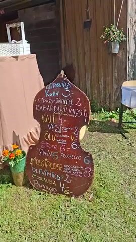The video opens with a close-up of a violin-shaped chalkboard menu displaying various drinks and food items with prices. The menu is leaning against a wooden structure. The camera then pans slightly to the left, revealing a table covered with a brown tablecloth, adorned with ruffles at the bottom. On the table, there are some items that are not clearly visible, and a woman in a green shirt is standing behind it. Further left, a green planter with orange flowers is placed on the ground.

The scene then shifts to show a tent with hanging string lights. Several people are standing in a line, seemingly waiting to be served at the table. The camera moves to show a wooden building in the background, with a stage set up in front of it, decorated with colorful vertical stripes and some white lace.

The camera then pans to the right, showing more of the wooden building and the surrounding grassy area. A few people are gathered near the entrance of the building. The video concludes with a wider shot of the outdoor area, showing more buildings with orange roofs and the sky. The overall atmosphere appears to be that of an outdoor market or event.