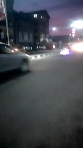 The video depicts a street scene at dusk or early evening. A silver SUV is initially visible in the foreground, parked on the side of the road. The street is illuminated by streetlights, with buildings lining the background. Cars are parked along the street. Subsequently, a man on a motorcycle drives past the camera, heading in the direction of the bright headlights in the distance. Finally, a white bus with its headlights on drives past, also moving away from the camera.