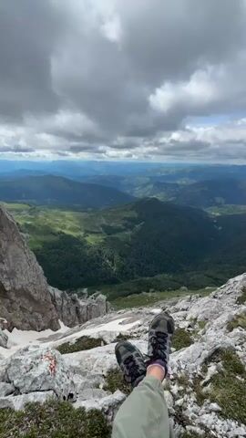 A first-person perspective shot from a mountaintop shows a person's legs and feet, clad in hiking boots, resting on rocks. The person is wearing green pants. The background reveals a vast mountain landscape with rolling green hills and valleys, under a cloudy sky. The camera is angled slightly downward, providing a view of the person's legs and then panning out to the expansive scenery. There is snow on the rocks in the foreground.