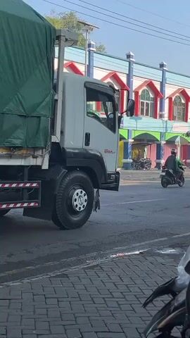 The video shows a white truck with a large green tarp covering its cargo. The truck is on a paved road. To the left of the truck, a person is riding a white motorcycle. In the background, there is a building with colorful trim and arched windows. The sky is visible and appears clear. The scene is outdoors during daylight.