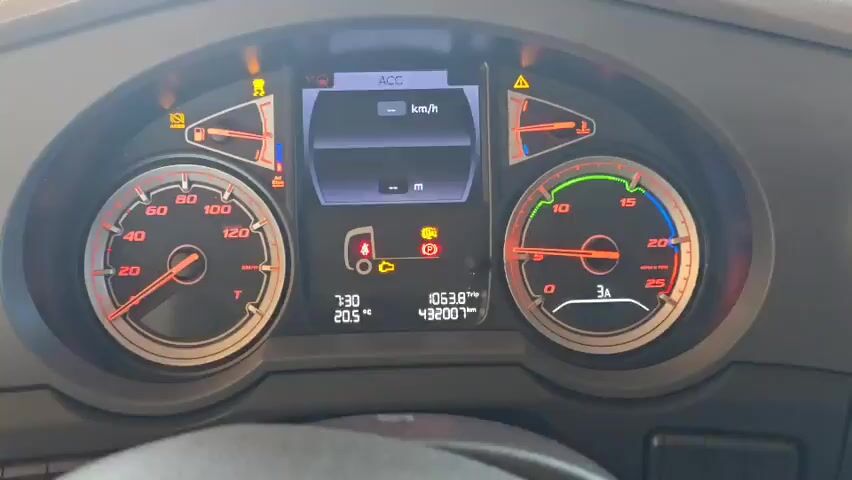 The video shows the dashboard of a vehicle, most likely a truck or a bus, while the engine is running. The dashboard features a speedometer on the left, a tachometer on the right, and a digital display in the center showing ACC (adaptive cruise control) information, time, temperature, and trip distance. Warning lights are illuminated, indicating possible issues with the vehicle. The tachometer needle fluctuates, corresponding to the engine idling. No people are visible in the frame.