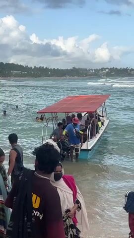 The video shows a light blue motorboat with a red canopy approaching a beach in a choppy sea. The boat is crowded with passengers, including a man in a white shirt standing on the edge of the boat. An individual in a blue shirt and pink hat stands in the water helping people disembark. Several people are standing on the beach, some wearing scarves or carrying bags. In the background, there is a coastline with buildings and trees. The boat is labeled “GLASS BOTTLE” and has a Sri Lankan phone number. People are wading in the water nearby. The sky is partly cloudy.