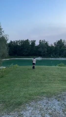 The media file depicts a person standing on a grassy area next to a body of water. The person is wearing a helmet, a light-colored t-shirt, and dark shorts, and appears to be on a unicycle. A backpack is slung over their shoulder.

The scene is set by a tranquil lake or river with lush green trees lining the opposite bank. The sky is a soft gradient of blue and purple, suggesting it is either dawn or dusk. The weather appears calm and pleasant. The ground in the foreground is covered in gravel, with a patch of well-maintained green grass where the person is positioned.

The overall mood is one of peacefulness and outdoor recreation. The person seems to be enjoying a moment of solitude or preparing for a ride. The location, Höslwang, Germany, provides a context of a natural, possibly rural or park-like environment. There is no visible text in the media file.