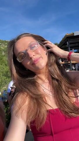 The video opens with a medium close-up of a young woman with long brown hair, wearing glasses and a reddish top. She is outdoors during the day, with a rocky, green hillside visible in the background. The camera then pans slightly, revealing other people behind her. One person is wearing a white hat and sunglasses, another is looking at their phone, and a third is partially visible. The lighting suggests a sunny day. The woman smiles and touches her hair as the video progresses. In the background, there is a wooden structure, possibly a deck or balcony, with some bottles and bags on it. The overall scene appears to be a casual outdoor gathering with a scenic backdrop.