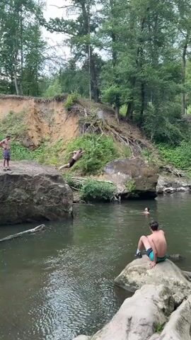 This is a video of a natural setting with a river flowing through it. The river has rocks and large boulders along its edges, and the surrounding landscape is lush with trees and greenery. The time of day appears to be daytime, with natural light illuminating the scene. The weather seems pleasant, possibly warm, given the activity taking place.

The main activity captured is people jumping into the river from a rocky overhang. In the first frame, a person is seen in mid-air, preparing to enter the water. The second frame shows the splash as they hit the water, creating a significant disturbance. Subsequent frames show the ripples and splashes subsiding, with other individuals already in the river. One person is sitting on a large rock in the foreground, facing away from the camera, observing the scene. Another person is visible in the water, swimming or wading.

The overall mood seems to be one of recreation and enjoyment in a natural environment. The setting suggests a popular spot for swimming and cliff jumping. The location is identified as Irrel, Germany, which is known for its scenic rivers and natural landscapes. There is no visible text in the video.