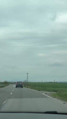 The video depicts a car driving on a paved road through a rural landscape. The road is straight and has white lines marking the lanes. Ahead, another car is visible, driving in the same direction. The surrounding environment is characterized by green fields extending towards the horizon. Power lines and towers line the edge of the field, adding to the industrial feel. The sky is overcast with grey clouds, casting a soft light over the scene. The video is taken from the perspective of someone inside a car, looking through the windshield. The dashboard is visible at the bottom of the frame.