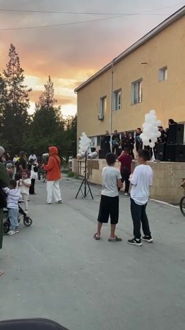 The video shows an outdoor gathering in front of a building with a light yellow facade. A group of people, possibly a band, is performing on a raised stage decorated with white balloons. The performers are dressed in black and appear to be singing and playing instruments. Several audience members are visible in the foreground, including children. Some children are looking towards the stage, while others are engaged in their own activities. The sky above is a gradient of orange, pink, and blue, indicating it is either sunrise or sunset. There are trees and outdoor lighting fixtures visible. The overall atmosphere appears festive and casual.