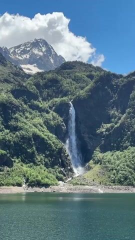The video shows a scenic view of a tall, narrow waterfall cascading down a lush, green mountainside into a calm lake or reservoir. The mountain behind the waterfall is partially covered in snow at its peak. The sky is blue with some white, fluffy clouds. The foreground is dominated by the water body, which appears green and placid. The scene is well-lit, suggesting a sunny day.