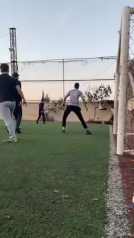Several people are playing soccer on an artificial turf field. A soccer net is visible on the right side of the frame. Players are dressed in athletic attire, and a person wearing gloves and a grey shirt stands near the goal. One person kicks a soccer ball toward the net. The players run across the field as the camera follows them. A tall metal structure is situated near the field.  The sky in the background is a light orange color, indicating it is late afternoon or early evening.