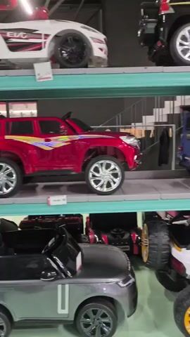 The video shows a multi-level storage rack filled with toy electric cars. The rack is made of green metal shelves supported by gray metal uprights. On the top shelf, there is a black toy SUV with colorful graphics and chrome wheels. Below that, on the second shelf, a red toy SUV is parked next to a blue toy Mercedes G-Wagon with red accents. On the third shelf, a white toy Mercedes G-Wagon is parked next to a black toy SUV. On the bottom shelf, a white toy off-road vehicle with large yellow and black tires is in the foreground, and a black and gold toy dune buggy is to its right. On the far right of the shelves, other toy cars are visible, including a green toy Mercedes G-Wagon, a red toy sports car, and a pink toy Mercedes G-Wagon. The environment appears to be an indoor storage facility or showroom, with exposed pipes and concrete walls visible in the background. There are no people visible in the video.
