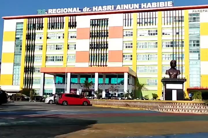 The video shows a frontal view of a multi-story hospital building with the name "RS. REGIONAL dr. HASRI AINUN HABIBIE" displayed prominently in large red letters across the top. The building has a bright, colorful facade with vertical yellow and white stripes on the sides and orange and white sections in the center. There are many windows on each floor. In front of the building is a paved area with several cars parked, including a prominent red car. A statue of a bust is located in the foreground, placed on a black and white pedestal. The sky is blue and clear. The overall impression is of a modern and well-maintained hospital.
