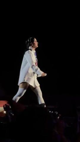 A person walks across a stage holding a microphone in their right hand. They are wearing a white oversized shirt, white thigh-high socks, and black shoes with buckles. The person has dark hair pulled back in a ponytail. The stage is dark with some colorful lighting in the background. The audience is visible in the foreground, with some hands raised.