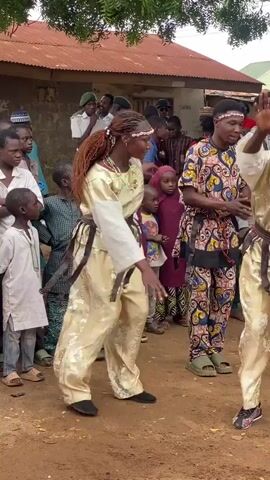 A group of people, including children and adults, are gathered outdoors in a dirt-floored area. Two young women are dancing in the foreground, dressed in traditional clothing. The woman on the left wears a gold outfit with brown fringe detailing around her waist and wrists, and a green beaded necklace. She has her hair braided with decorative bands. The woman on the right wears a patterned shirt and pants with colorful designs. Both dancers appear to be energetically moving and smiling. The crowd watches them, some with their hands raised. Behind the dancers, several men in uniform, possibly security personnel, stand near a building with a corrugated metal roof. The environment suggests a rural or semi-urban setting, possibly during a cultural event or celebration.