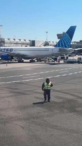 A person dressed in a high visibility vest and protective face covering stands on an airport tarmac, holding a red signaling wand. Behind them, a United Airlines airplane is parked at an airport gate. The plane's blue tail and UNITED lettering are clearly visible. Various airport vehicles and equipment are scattered around the aircraft. The day appears sunny and clear.