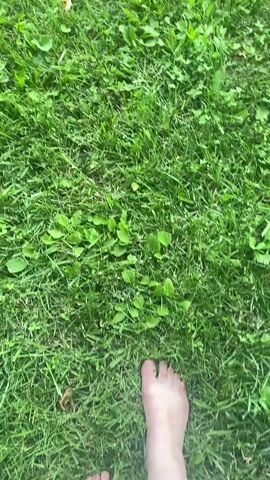 The video shows a first-person perspective of someone walking barefoot on green grass. The grass is well-maintained, with some small, broad-leafed plants interspersed among the blades. A single dry, brown leaf is visible on the grass in the upper left quadrant of the frame. The shot is tightly focused on the ground and the person's feet, suggesting an informal and relaxed activity. The lighting suggests it is daytime, likely with overcast or diffused sunlight, as there are no harsh shadows. The scene appears to be outdoors, possibly a park, a lawn, or a backyard. The overall mood is one of simple, grounded connection with nature. The location is Târgu Neamț, Romania. There is no visible text in the video.