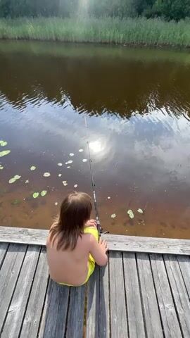 A person with long brown hair, wearing yellow shorts, sits on a wooden dock and fishes in a body of water. The sun is visible reflecting on the water, and there are reeds and trees in the background. The camera pans slightly to the right.