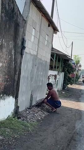 A man, shirtless and wearing dark shorts, squats on the dirt beside a partially constructed building and works on the base of the wall with his hands. A pile of rubble is next to him. The building appears to be made of cinder blocks and is covered with a rough layer of gray material on its side. A pipe extends from the building's wall. Across a paved road, several people on motorcycles are seen moving away from the camera, and a woman in a long gray dress and blue headscarf walks toward the right of the frame, carrying a broom and dustpan. The environment is outdoors under a clear sky, with power lines visible overhead and trees and other buildings in the background.