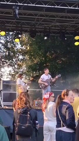 A live outdoor concert is underway on a grassy area. A band of at least three musicians is performing on a stage. The band members are playing guitars and singing. The stage is lit by overhead stage lights. A crowd of people are watching the performance from the grass in front of the stage. Some people in the crowd are holding drinks or phones. The sun appears to be setting, casting a warm light on the scene. The background is filled with trees.