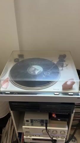 This is a close-up, eye-level shot of a white turntable sitting on top of a white shelf unit. A vinyl record is on the turntable. Beneath the turntable, on the first shelf of the unit, is a black stereo component. On the second shelf are two silver stereo components. On the third shelf are two more silver stereo components, and on the bottom shelf, leaning against the unit, is a record album with a colorful abstract design. To the right of the shelf unit, headphones are resting on top of a stack of CDs, and more CDs are piled on the floor. Several CDs are also stacked on the far left of the image. The background is a plain wall, and a window is visible at the top of the frame.