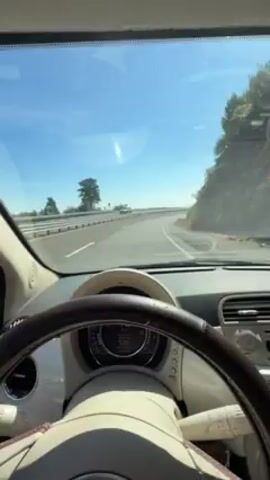 The video shows a driver's point of view from inside a light-colored Fiat 500 while driving on a winding highway during a sunny day. The road is lined with a metal guardrail on the left and a steep, vegetated hillside on the right. Other cars can be seen in the distance on the highway. The dashboard and steering wheel are visible in the foreground.