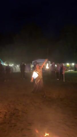 A fire dancer in a dark-colored outfit with gold embellishments performs with two flaming props in a low-lit outdoor area at night. The dancer, with long dark hair, moves gracefully, creating fiery circles and patterns. In the background, several people are watching, and there are white tents and string lights, suggesting a festive or communal event. The ground appears to be dirt or sand, illuminated by the fire.