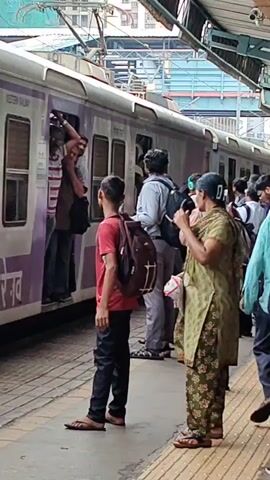 The video shows a busy train station platform with a train stopped at the platform. Several people are standing on the platform, some appearing to be waiting to board the train, while others are already inside. The train is purple and white with "WR" visible on its side, and what appears to be "PASCHIM RAILWAY" written in purple lettering. People are dressed in casual clothing, and some are carrying bags. The platform has yellow tactile paving. The atmosphere appears to be one of daily commuter activity.
