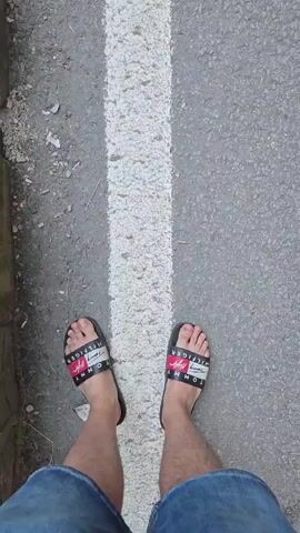 The video begins with a close-up shot of a person's feet wearing black slides with red and white accents as they walk on a paved road with a white line. The camera pans up slightly to reveal the person's legs and the surrounding environment, which appears to be a roadside with rocks on the left. The video then transitions to a wider shot, showing the road curving alongside a rocky cliff face. There are several vehicles on the road, including a white van and a yellow truck. People are seen sitting and standing near the roadside. The camera then tilts upwards, showing the immense scale of the rocky cliff and a distant mountain covered in trees. The video captures the progression of walking along a road bordered by natural rock formations and mentions a body of water on the left.