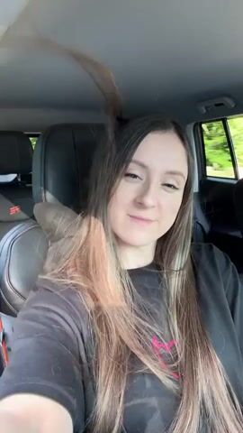 A woman is sitting in the passenger seat of a car, looking towards the camera. She has long, wavy brown hair with blonde highlights and is wearing a black t-shirt with a pink design. The car's interior is visible, including a black leather seat with red stitching. Outside the car, trees and greenery can be seen through the window, suggesting the car is in motion and driving through a wooded area.