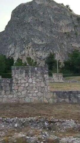 The video shows a drone shot panning across a rocky mountainside, with a stone wall in the foreground. There is a playground structure visible behind the wall, as well as some trees and a building on the hillside. The sky is overcast.