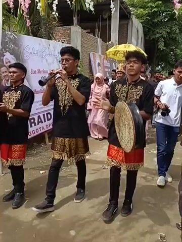 Two young men are walking in the foreground, one playing a flute and the other a drum. They are dressed in traditional black and gold clothing with red accents. Behind them, a wedding procession is taking place. A groom, dressed in an ornate traditional suit with a gold and red patterned jacket and a matching hat, is being escorted by two women. He is holding hands with one woman on his left and the other on his right. A yellow umbrella is being held over the groom's head. Several other people are walking in the procession behind them. The environment appears to be outdoors, possibly in a village or a residential area, with a banner in the background that seems to be for a wedding or an event. The ground is unpaved.