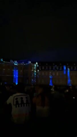 The video shows a nighttime outdoor scene with a crowd of people in the foreground. They are facing a large building illuminated with blue lights and projections. Blue light beams are visible projecting from the building into the dark sky. The building appears to be an ornate structure with multiple windows. The crowd is silhouetted against the light, and some individuals are wearing white shirts with graphic designs. The overall atmosphere suggests an event or performance taking place in front of the illuminated building.
