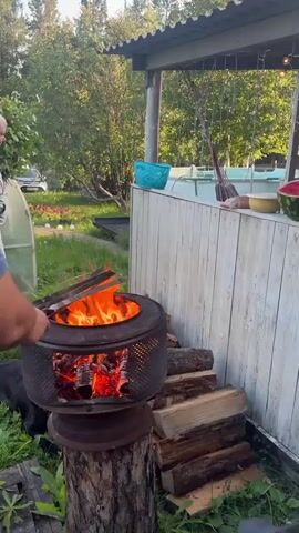 In an outdoor setting, a man in a blue t-shirt and dark shorts is tending to a fire in a makeshift grill. The grill appears to be made from the drum of a washing machine, which is placed on top of a tree stump. A pile of chopped firewood is stacked to the right of the grill. In the background, an older man in a gray tracksuit walks away from the camera, and a greenhouse is visible on the left. The overall environment suggests a casual, perhaps rural, gathering.