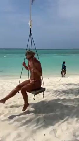 A young woman in a pink bikini is sitting on a wooden swing suspended from a palm tree at the edge of a white sand beach. She is blonde and is wearing sunglasses. The woman is smiling and talking, looking directly at the camera. Behind her, there is a shallow, turquoise-colored ocean under a partly cloudy blue sky. There are other people in the water and walking along the shoreline in the background. The beach has shadows cast by the tree. The swing is stationary.